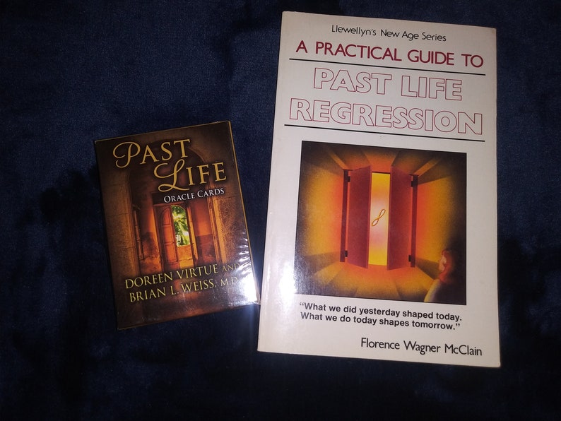 Puede incluir: Un libro titulado "A Practical Guide to Past Life Regression" de Florence Wagner McClain, parte de la serie New Age de Llewellyn. La portada del libro presenta una puerta con una luz brillante que brilla a trav&eacute;s. Junto al libro hay una baraja de cartas de or&aacute;culo titulada "Past Life Oracle Cards" de Doreen Virtue y Brian L. Weiss, M.D.