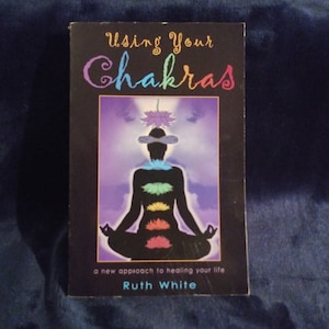 May include: Book cover with a purple background and the title "Using Your Chakras" in a white cursive font. The cover features a silhouette of a person sitting in a lotus position with seven colorful chakras along the spine. The text "a new approach to healing your life" and the author's name "Ruth White" are printed below the image.