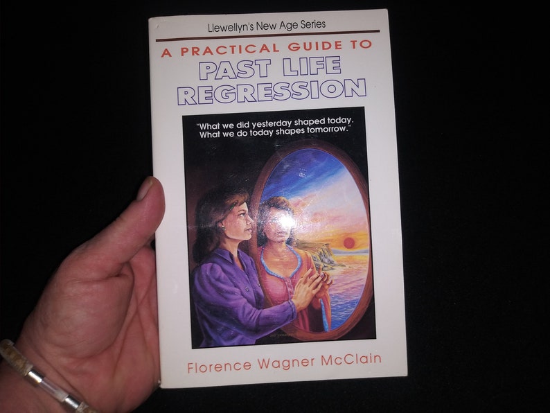 May include: A white book with a blue and purple illustration of two women looking at each other in a mirror. The book title is "A Practical Guide to Past Life Regression" and the author is Florence Wagner McClain. The book is part of Llewellyn's New Age Series.