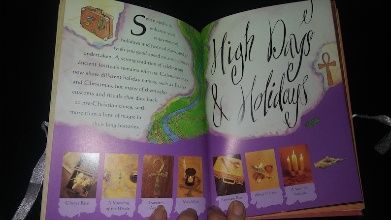 May include: An open book with a purple cover and a title that reads "High Days & Holidays". The book features illustrations of various holiday-themed items, including a suitcase, a whale, a traveler's amulet, a wish, a sandan vase, wheat husks, and candles.