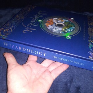 May include: A blue hardback book titled "Wizardology: The Secrets of Merlin." The cover has gold foil detailing and a circular design with illustrations. The book is held by a hand.