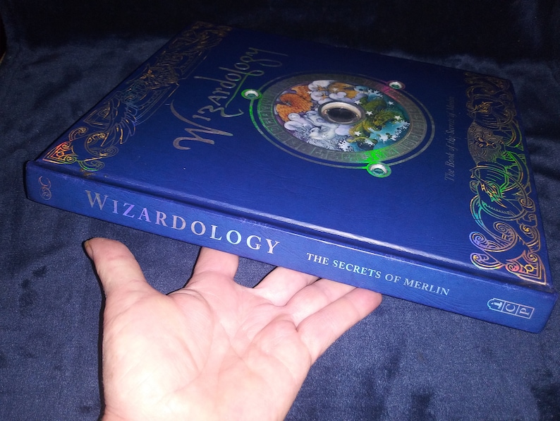 May include: A blue hardback book titled "Wizardology: The Secrets of Merlin." The cover has gold foil detailing and a circular design with illustrations. The book is held by a hand.