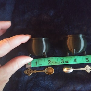 May include: Two small, black cauldron-shaped containers with three legs, next to a measuring tape. The cauldron is approximately 2 inches in diameter. Two decorative spoons are also present, one gold and one silver.