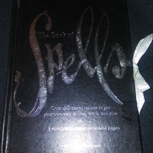 May include: A black book with a silver title that reads "The Book of Spells". The book has a silver inscription that reads "Over 40 secret recipes to get your own way in love, work, and play. 3 chapters with our sealed pages."