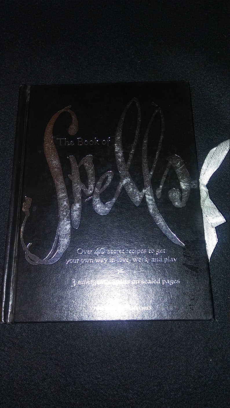 May include: A black book with a silver title that reads "The Book of Spells". The book has a silver inscription that reads "Over 40 secret recipes to get your own way in love, work, and play. 3 chapters with our sealed pages."