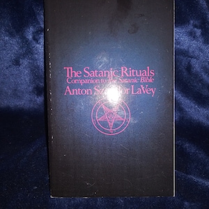 May include: A black book cover titled The Satanic Rituals, Companion to The Satanic Bible by Anton Szandor LaVey. The text is printed in magenta, and a magenta pentagram inside a circle is centered below the author's name on the front cover.