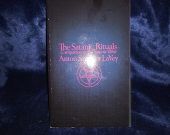 May include: A black book cover titled The Satanic Rituals, Companion to The Satanic Bible by Anton Szandor LaVey. The text is printed in magenta, and a magenta pentagram inside a circle is centered below the author's name on the front cover.