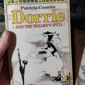 May include: A book cover with a yellow title "Dorrie" and the subtitle "And The Wizard's Spell". The cover features a black and white illustration of a girl with a black hat and a black cat.