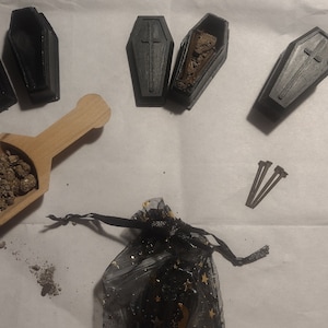 May include: A set of miniature black coffin-shaped boxes with a cross design, filled with brown dirt. A wooden scoop with brown dirt, three silver nails, and a black mesh bag with gold stars are also included.