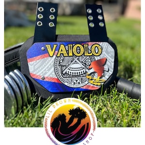 May include: A black sports chest protector featuring a central plate with the name VAIOLO in yellow text, a graphic of a red eagle, and a circular emblem with Samoan patterns. The plate is attached to black straps with silver metal eyelets.