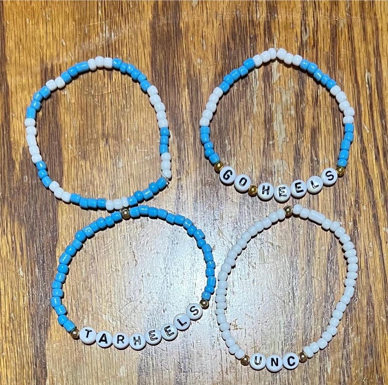 Op de afbeelding: Vier armbanden met kralen. Twee armbanden zijn blauw en wit, &eacute;&eacute;n zegt "GO HEELS" en de andere "TARHEELS". De andere twee armbanden zijn wit en zeggen "UNC".