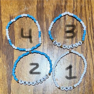 Op de afbeelding: Vier armbanden met kralen. De armbanden zijn gemaakt van kleine, ronde kralen in wit en lichtblauw. E&eacute;n armband spelt "UNC", een andere "TARHEELS" en een andere "GO HEELS". De vierde armband heeft het nummer "4".