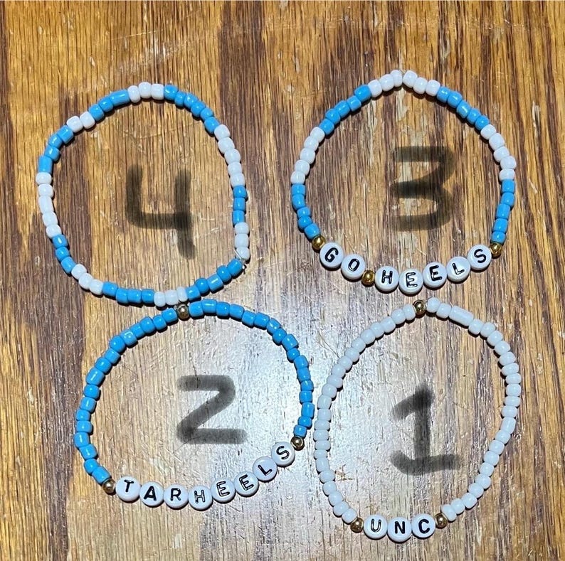 Op de afbeelding: Vier armbanden met kralen. De armbanden zijn gemaakt van kleine, ronde kralen in wit en lichtblauw. E&eacute;n armband spelt "UNC", een andere "TARHEELS" en een andere "GO HEELS". De vierde armband heeft het nummer "4".