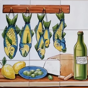May include: A decorative tile mural depicting a kitchen scene. It features hanging fish, a wine bottle with a label, cheese, olives, lemons, and a decorative plate. The color palette includes blue, yellow, green, and brown.