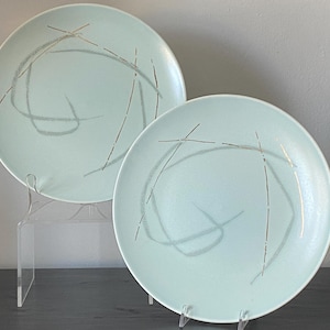 May include: Two pale blue ceramic plates with a mid-century modern design. Each plate has a grey and gold abstract pattern. The plates are displayed on clear acrylic stands, highlighting their design.