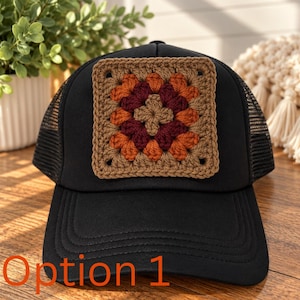 May include: A black trucker hat featuring a square crochet patch on the front panel. The patch is crafted in a granny square pattern using yarn in shades of brown, dark burgundy, and burnt orange. The hat has a solid black brim and mesh back.
