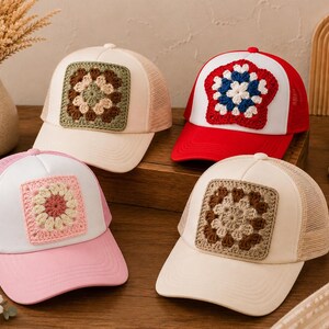 May include: Four trucker hats featuring crocheted granny square patches on the front panels. One hat is cream with a green and brown patch, one is red and white with a red, white, and blue patch, one is pink and white with a pink and cream patch, and one is cream with a brown and cream patch.