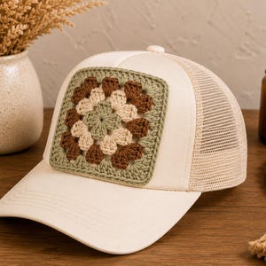 May include: Cream-colored trucker hat with a crocheted square patch. The patch features a floral design in shades of brown, beige, and green. The hat has a mesh back and a curved brim, and a button on top.
