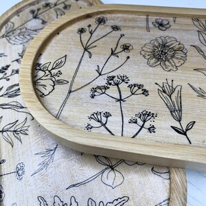 May include: Two round wooden trays with a floral design. The trays are made of light brown wood and have a white background. The floral design is black and features various flowers and leaves.