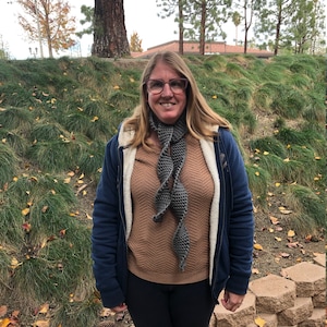 Może przedstawiać: Kobieta w niebieskiej kurtce, brązowym swetrze, czarnych spodniach i szarym dzianym szaliku. Stoi przed ceglaną ścianą i zieloną trawą.