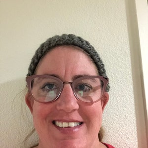 Może przedstawiać: Kobieta z ciepłym uśmiechem ma na sobie szary dzianinowy opaska na głowę i różowe okulary typu „kocie oko”. Ma na sobie czerwoną koszulę.