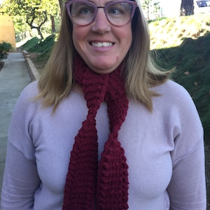 Może przedstawiać: Kobieta w jasno-fioletowym swetrze i bordowym dzianym szaliku. Szalik jest luźno zawiązany wokół szyi.