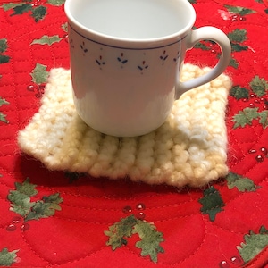 Może przedstawiać: Biały ceramiczny kubek z niebieskim i czerwonym wzorem kwiatowym stoi na kremowym dzianym podkładce. Podkładka leży na czerwonej pikowanej powierzchni z zielonym wzorem liści ostrokrzewu.