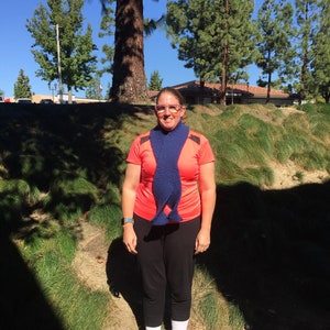 Może przedstawiać: Osoba w różowej koszuli i niebieskim dzianym szaliku. Osoba stoi przed trawiastym terenem z drzewami w tle.