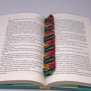 Może przedstawiać: Zbliżenie na zakładkę wykonaną na szydełku w tęczowych kolorach, w tym różowym, zielonym, żółtym i fioletowym. Zakładka znajduje się w otwartej książce z tekstem o planowaniu wydarzeń i kalendarzu.