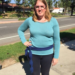 Może przedstawiać: Kobieta w turkusowym swetrze z niebieskim paskiem z dzianiny. Pasek jest zawiązany z przodu w supeł, a końce zwisają.