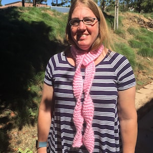 Może przedstawiać: Kobieta w pasiastych sukienkach i różowym dzianym szaliku. Uśmiecha się i patrzy w obiektyw.