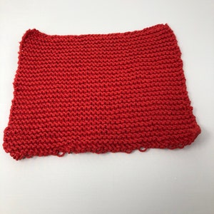 Może przedstawiać: Czerwona dziana ściereczka do naczyń lub gąbka. Ściereczka jest wykonana z grubego, fakturowanego włókna i ma lekko strzępiony brzeg.