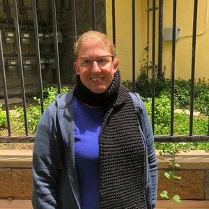 Może przedstawiać: Kobieta w niebieskiej kurtce, niebieskiej koszuli i czarnym dzianym szaliku. Stoi przed czarnym metalowym ogrodzeniem.