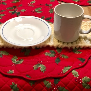 Może przedstawiać: Biały ceramiczny kubek z uchwytem stoi na białym dzianym podkładce. Podkładka leży na czerwonym pikowanym podkładce ze wzorem ostrokrzewu. Biały ceramiczny spodek leży na podkładce.