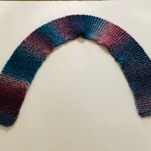 Może przedstawiać: Wełniany szalik w odcieniach fioletu, niebieskiego i turkusowego. Szalik jest złożony na pół i leży płasko na białym tle.