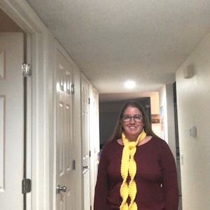 Może przedstawiać: Kobieta w bordowym swetrze i czarnych spodniach stoi na korytarzu. Ma na szyi żółty, szydełkowy szalik.