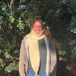 Może przedstawiać: Kobieta w białym dzianym szaliku, szarej kurtce i niebieskich dżinsach. Stoi przed zielonym krzewem.