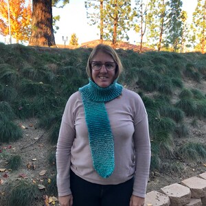 Może przedstawiać: Turkusowo-niebieski dziergany szalik typu cowl. Szalik jest noszony wokół szyi i zwisa z przodu. Tło obejmuje zieloną trawę, drzewa i błękitne niebo.