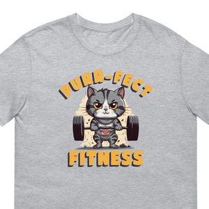 Może przedstawiać: Szara koszulka z nadrukiem kreskówkowego kota podnoszącego ciężary. Na koszulce widnieje napis "PURR-FECT FITNESS".