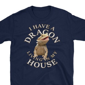 Può includere: Una t-shirt blu navy con una grafica circolare bianca che dice "Ho un drago che vive nella mia casa". Un drago barbuto è raffigurato all'interno del cerchio.