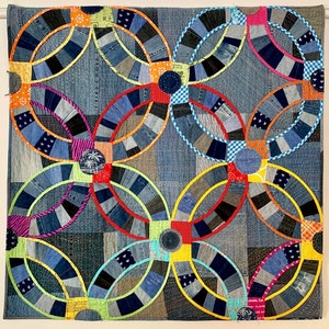 Könnte beinhalten: Handgefertigte Steppdecke mit floralem Muster. Sie besteht aus recyceltem Denim in verschiedenen Blautönen, mit farbenfrohen Akzenten in Orange, Rot, Gelb und Grün. Das Design zeigt ineinandergreifende Kreise, die ein einzigartiges Textil bilden.