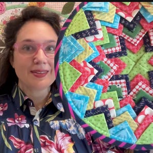 May include: A vibrant, handmade quilt with a geometric pattern. The quilt incorporates various fabrics in blue, green, red, and yellow. The quilt is partially visible, with a woman wearing glasses and a floral shirt in the background.