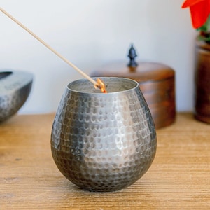 May include: A silver hammered metal candle holder with a wooden stick burning inside. The candle holder is on a wooden surface.