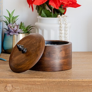 May include: A wooden box with a lid, the lid is open and the box is on a wooden surface. There is a book, a potted plant, a vase with a red poinsettia, and a white ceramic candle holder with a gold top on the surface.