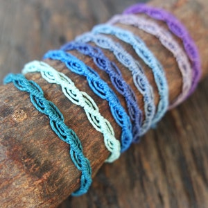 Può includere: Un set di otto braccialetti all'uncinetto colorati in tonalità di blu, verde e viola. I braccialetti sono disposti a forma di arcobaleno e sono esposti su una superficie di legno.