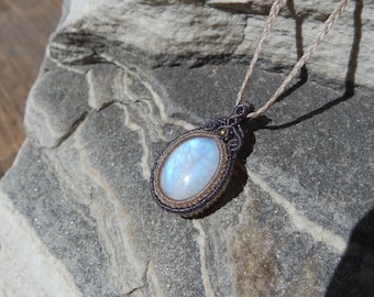 Puede incluir: Un colgante con una piedra de luna ovalada de brillo azulado, engastada en un intrincado nudo de macramé con cordones de color marrón oscuro y beige. El colgante cuelga de un cordón trenzado fino de color marrón claro, mostrando detalles.