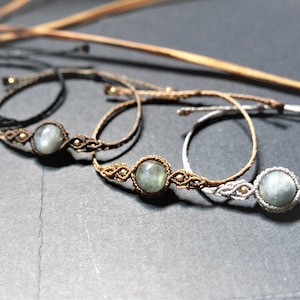 Peut inclure: Quatre bracelets en macramé avec une pierre précieuse grise, ronde et centrale. Les bracelets varient en couleur, notamment noir, marron et blanc, avec des nœuds complexes et de petites perles dorées. Ils sont présentés sur une surface gris foncé.