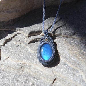 Puede incluir: Un collar con colgante ovalado azul con un cordón gris tejido. El colgante tiene una piedra azul engastada en un marco gris tejido con detalles dorados. El collar se muestra sobre una superficie de piedra gris.