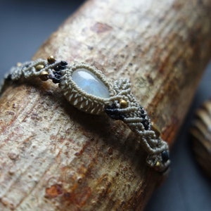 May include: A white macrame bracelet with a large oval moonstone bead and gold accents. The bracelet is wrapped around a piece of driftwood.