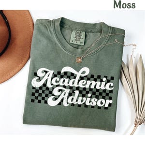 May include: A moss green t-shirt with a white retro-style graphic that says "Academic Advisor" with a black and white checkered pattern behind the text.